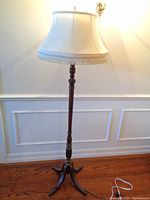 Full view of vintage wooden floor lamp with white fabric lampshade with fringe trim illuminated