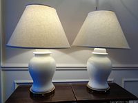 Pair of white ceramic table lamps with beige shades shown side by side on wooden surfaces against a white wall with wainscoting.