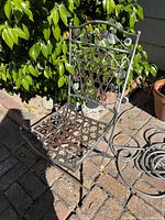 One metal folding chair with decorative leaf and vine detailing on the backrest and seat, photographed outdoors on a stone surface beside plants.