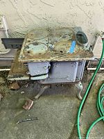 Cast iron child's toy stove with four circular burner plates and an oven door, showing rust and wear outdoors on concrete ground near a wall.