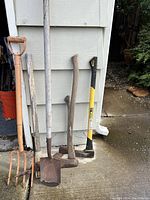 Photo showing six garden tools including an axe, splitting maul, digging shovel, post hole digger, garden fork, and another wooden handled tool leaning against a wall.