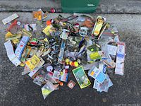 Wide image showing an assortment of fishing gear including lures, hooks, bobbers, weights, and packaged items laid out on a concrete surface.