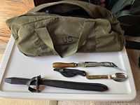 US military olive green duffel bag with camping gear including bayonet, folding knife, and camp spoon/fork on tray