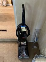 Full view of black Bissell ProHeat upright carpet shampooing machine standing on carpet. Machine has coiled hose attached and power cord wrapped on side.