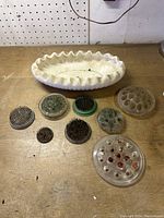 Photo showing one oval white ceramic vase with ruffled edge on wood surface alongside eight assorted flower frogs made of metal, glass, and ceramic.