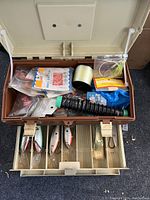 Open fishing tackle box showing top and bottom compartments filled with fishing lures, fishing line spool, and terminal tackle items.