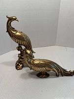 Two golden bird figurines shown, one standing upright with detailed feathers and another reclining with a long tail on a neutral backdrop.