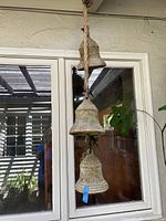 Three large metal bells hanging vertically on a thick rope, shown against a glass window reflection. Bells are weathered with patina.