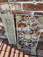 Photo showing two outdoor decorative wall plaques, one metal with scrollwork and gold accents, one stone or concrete with raised floral design, placed against a brick wall