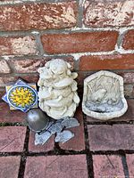 Four small garden decoration items including a sun face tile, Buddha figure, bird bath or feeder, and metal heart wind chimes placed against a brick wall on brick flooring.