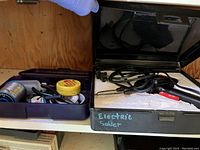 Soldering iron inside black plastic case marked 'Electric Solder' with visible soldering iron cord and tip.