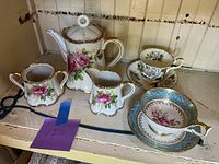 Main photo of the tea set including teapot, sugar bowl, creamer, and two cups with saucers, with floral patterns and gold accents.