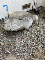 Photo of weathered concrete turtle hose holder placed on gravel by a building wall.