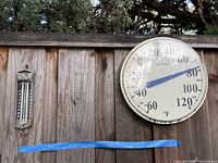 Wide shot of two thermometers mounted on wooden fence: large round La Crosse thermometer and rectangular Taylor thermometer.