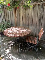 View of round wood slat table with metal base and one matching chair beside it, showing natural outdoor setting.