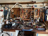 Full view of workbench with assorted tools hung on pegboard and placed on bench surface showing a variety of hand tools, clamps, containers, and miscellaneous items.