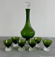 Front view of emerald green glass decanter with stopper and six matching green glasses with ribbed clear bases arranged in a semi-circle