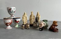 Photo of the full collection showing three resin statues of Chinese gods on round wooden bases, three hand-painted floral and bird motif egg cups, green pottery piece, brown dog figurine, and two character head ceramic figures.