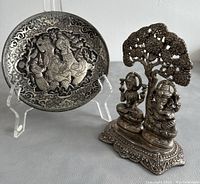 Photo showing a hand engraved Persian metal plate on display stand alongside the metal figurine of Lakshmi and Ganesha under a tree.