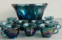 Front view of the blue carnival glass punch bowl surrounded by all 12 matching cups showing grape and vine pattern and iridescent finish