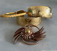 Overview of the four jewelry pieces including three gold-tone cuff bangles and one brooch on a grey textured surface.