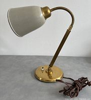 Side view of Mid-Century Modern brass desk lamp showing white conical metal shade, brass adjustable arm with gooseneck, round brass base with toggle on/off switch and brown power cord coiled beside it.