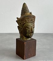 Front left diagonal view of vintage Thai metal Buddha head with detailed headdress, mounted on dark wooden base.