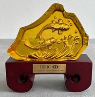 Front view of amber-colored Wedgwood glass art sculpture with embossed fish and water design placed on dark wooden stand. Metal plaque on stand shows edition number 18483 of 38888, and HSBC logo.