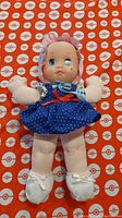 Front view of the 1980s nursery doll with blue eyes, wearing a blue polka dot dress with a red bow, white socks, and a bonnet.