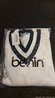 The new folded white Benin sweater with navy V-neck visible, in plastic packaging, on a dark surface.