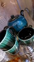 Photo showing teapot, partially obscured cups in front with blue-green glaze reflecting light.