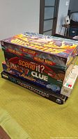 Stacked boxed board games on table including Battleship, Scene It?, Clue, Disney Aladdin, and Sports Battleship