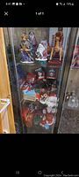 Three glass shelves filled with assorted Christmas holiday figurines, ornaments, and decorations including various Santa Claus figures, a snowman figure, a small wooden rocking horse, and holiday-themed collectibles.