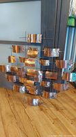 Front view of an abstract copper wall decoration sculpture with curved copper plates mounted on metal rods in geometric pattern on wooden floor.