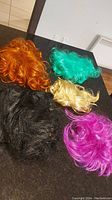 Photo showing all five wigs in orange, teal, blonde, black, and pink colors arranged on a dark surface