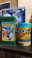 Two tins on shelf, one rectangular green The Jungle Book tin and one cylindrical Looney Tunes tin