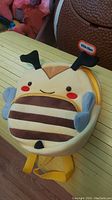 Front view of the bee-shaped kids backpack showing smiling face, antennae, and wings, placed on a wooden floor with other toys visible nearby.