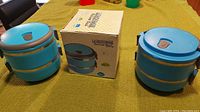 Two blue lunchboxes with two compartments each placed on a table with original packaging box behind them.