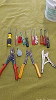 Various hand tools laid out on a green fabric, including multiple screwdrivers with different colored handles, wire cutters, locking pliers, and a small pulley.