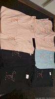 Eight t-shirts arranged flat, showing pastel pink, black, and light blue colors with moose embroidery