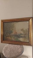 Photo of framed landscape painting showing church steeple by water and trees in a gold-colored frame
