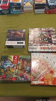 Four boxed jigsaw puzzles, laid out on a green surface including scenic and themed puzzles.