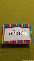 Top of game tin box showing the "What? Deluxe Edition" label with colorful stripes.