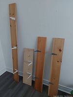 Four wooden shelves of varying lengths leaning against a white wall on a dark floor, each with metal brackets attached.
