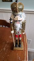Front view of 16-inch nutcracker with gold sequined crown, white beard, and holding staff on a gold base.