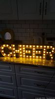 Lighted Cheer sign spelling 'CHEER' lit with warm white bulbs on a kitchen counter with drawers and tiled wall background.