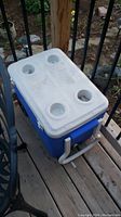 Blue and white Coleman wheeled cooler with four cup holders on lid, pull handle, and wheels, shown outdoors on wooden deck.