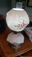 Side view of the vintage electric hurricane lamp showing frosted white glass globe with embossed floral and ribbon designs and pink flower painting, mounted on glass base with matching decoration and ornate metal stand.