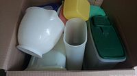 Box containing multiple vintage and modern Tupperware containers in various shapes and colors including green, yellow, pink, white, and teal, showing general contents of lot.