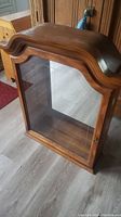 Front view of wooden wall cabinet showing curved top molding and glass door with wooden frame.